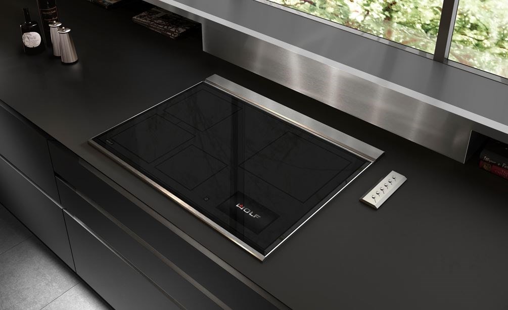 La surface de cuisson à induction Wolf 30 po de style transitionnel (CI30460T/S) est présentée dans un design de cuisine ultra-moderne élégant avec des plans de travail noirs lisses, un dosseret en acier inoxydable et des armoires sans poignée 